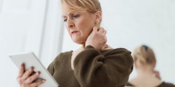 Contemplative mature female in casual wear watching video on tablet and touching neck while standing against mirror in light room