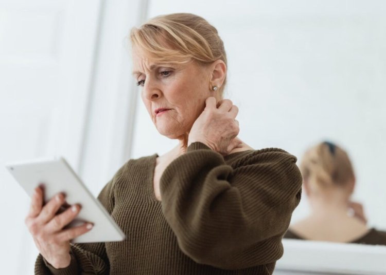 Contemplative mature female in casual wear watching video on tablet and touching neck while standing against mirror in light room