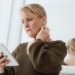 Contemplative mature female in casual wear watching video on tablet and touching neck while standing against mirror in light room